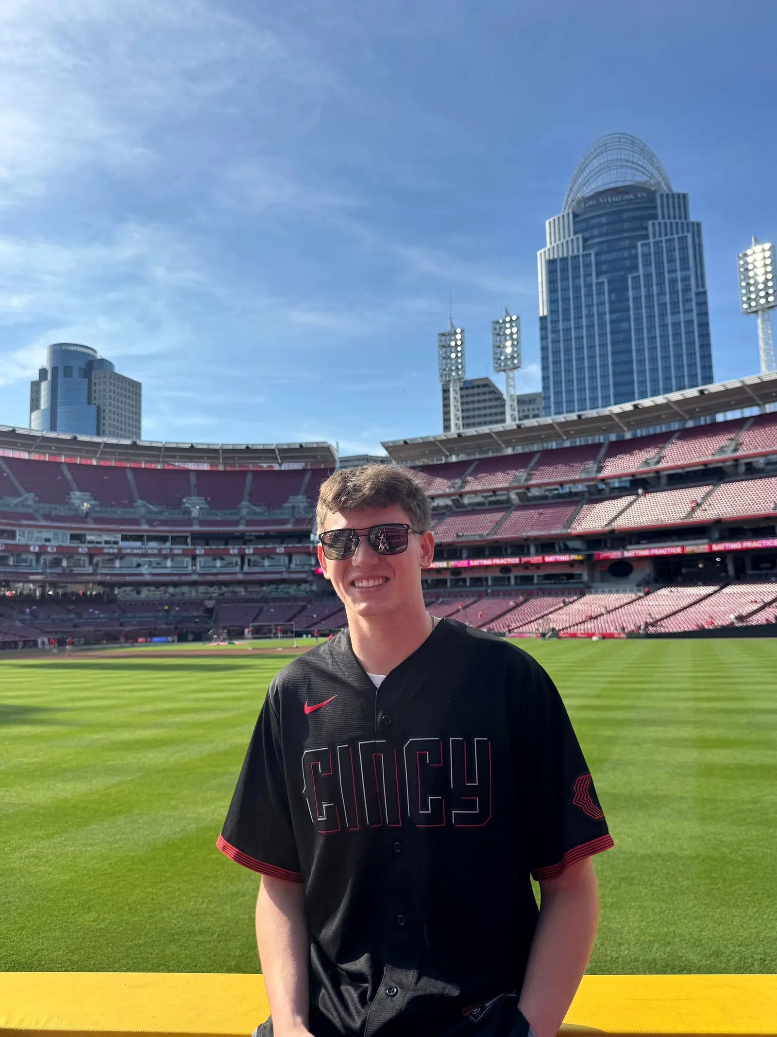 Nick Bibelhausen on the field at the Cincinnati Reds' ballpark, wearing a Cincy jersey and sunglasses, with the Great American Tower and the downtown skyline rising behind the stands.