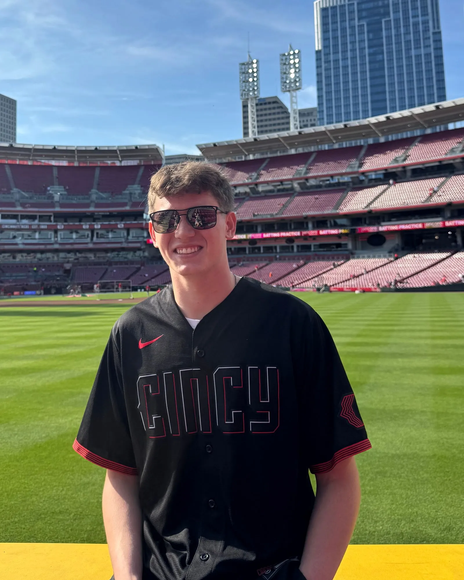 Nick Bibelhausen, founder of Bibelhausen Digital, standing on the field at the Cincinnati Reds' ballpark in a Cincy jersey and sunglasses, with the downtown skyline behind him.
